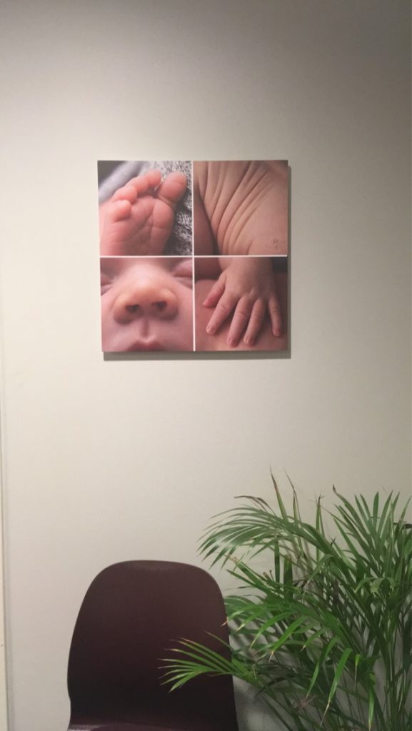 Et sammensatt bilde av babydetaljer henger på veggen over en stol og en plante på venterommet. 