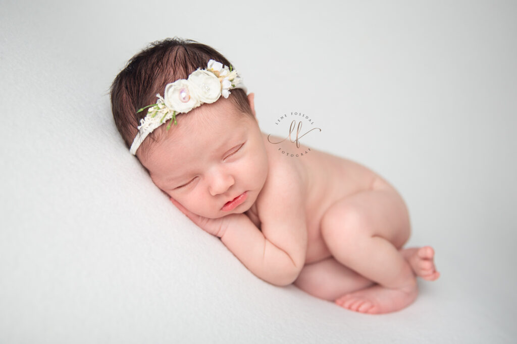 En nyfødt jentebaby som ligger på hvit bakgrunn. Hun har blomsterhårbånd på hodet og et lite smil på leppene.