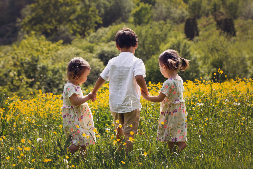 Tre barn med ryggen til i en eng av gule blomster. 