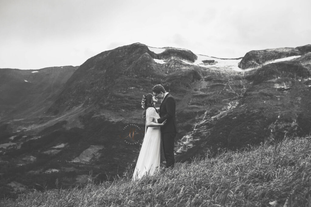 Et brudepar som står i en skrå eng og kysser. Bak seg har de fjelltopper med snø på. 