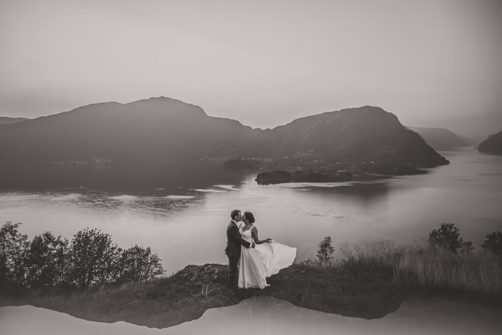 Et svart og hvitt bilde av et brudepar med en fjord og fjell bak seg. 