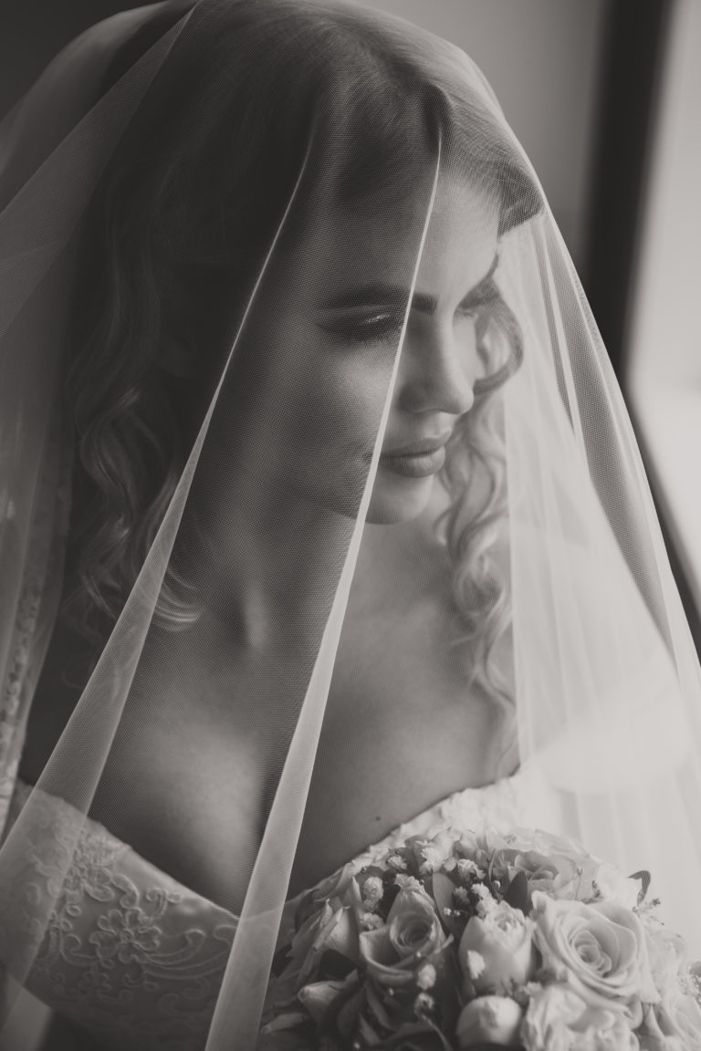 Dette bildet er av en brud under sløret. Det er et nærbilde, og hun ser mot høyre i bildet, ut vinduet. Vi ser toppen av brudebuketten som hun hoder.