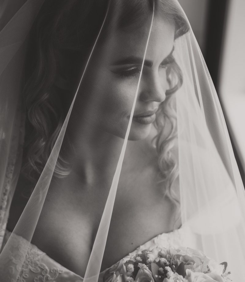 Dette bildet er av en brud under sløret. Det er et nærbilde, og hun ser mot høyre i bildet, ut vinduet. Vi ser toppen av brudebuketten som hun hoder.