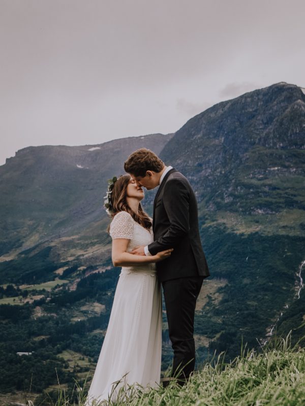 Sophie og Kjetil (brudepar) står i en bratt eng av grønt gress og nesten kysser. Bak dem er det høye fjell. Sophie har på blomsterkrans.