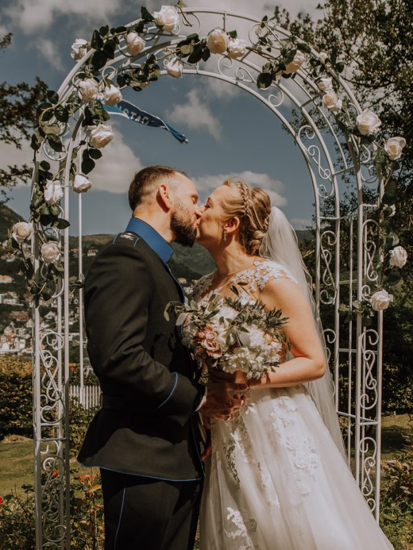 Bilde av Remie og Marta Elise som gir hverandre sitt første kyss som mann og kone. De er ute, de står under en blomsterbue og det er strålende sol. Remie har på seg uniform, Marta Elise har brudekjole.