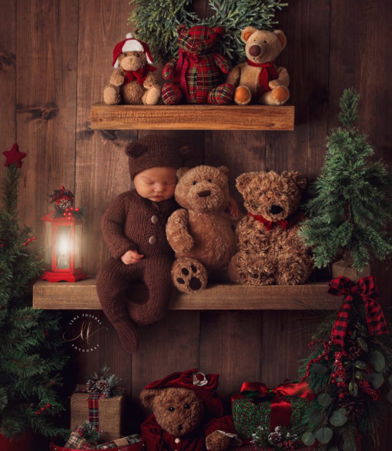 Bildet er av en baby som ser ut til å sitte sovende på en hylle sammen med masse bamser. Det er juletema på oppsettet. Babyen har på seg brun drakt og lue med bamseører.